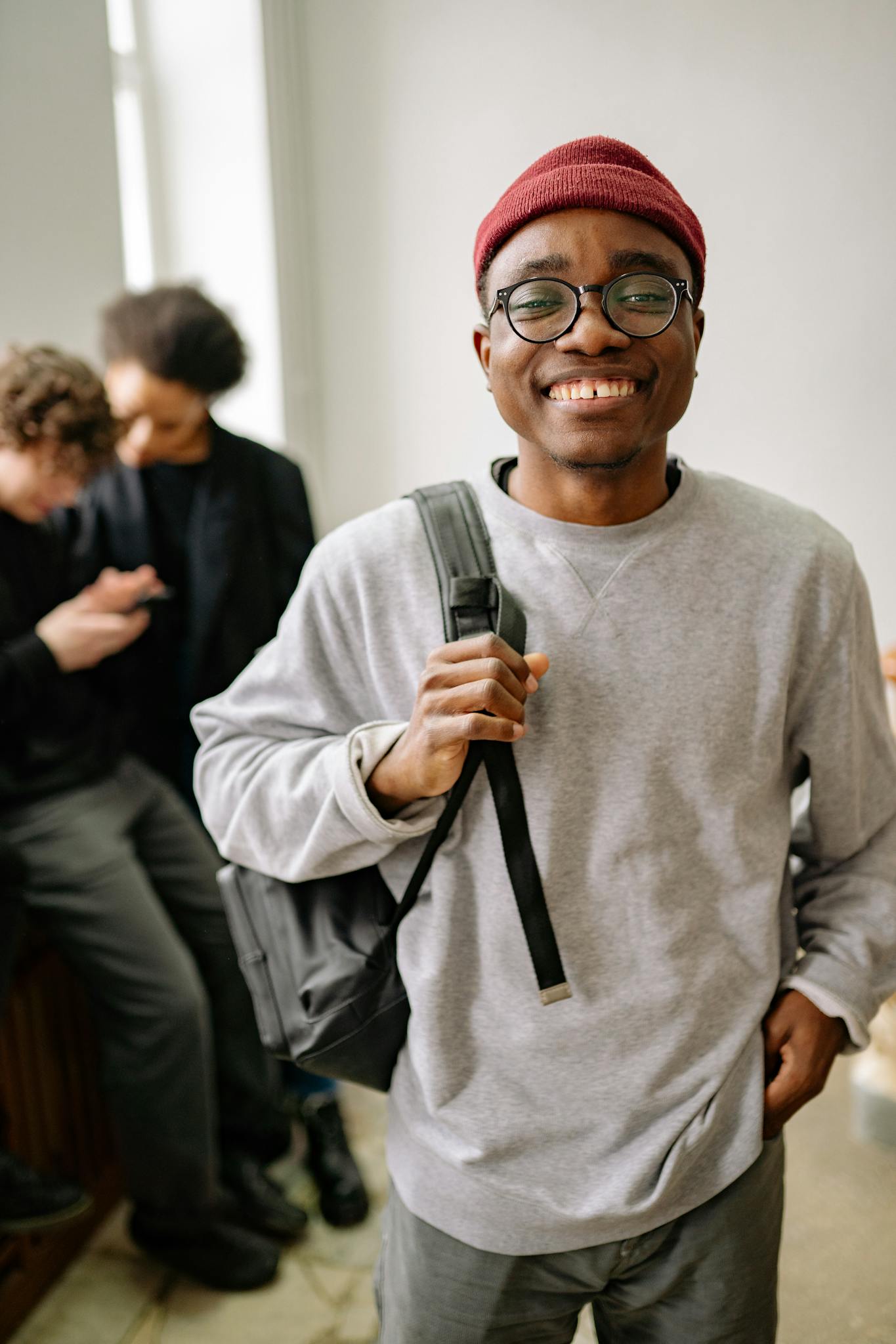 A cheerful young man in a gray sweater and glasses, smiling indoors with friends in the background.