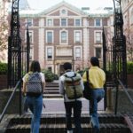 A group of students with backpacks walks towards a university building through an arch.