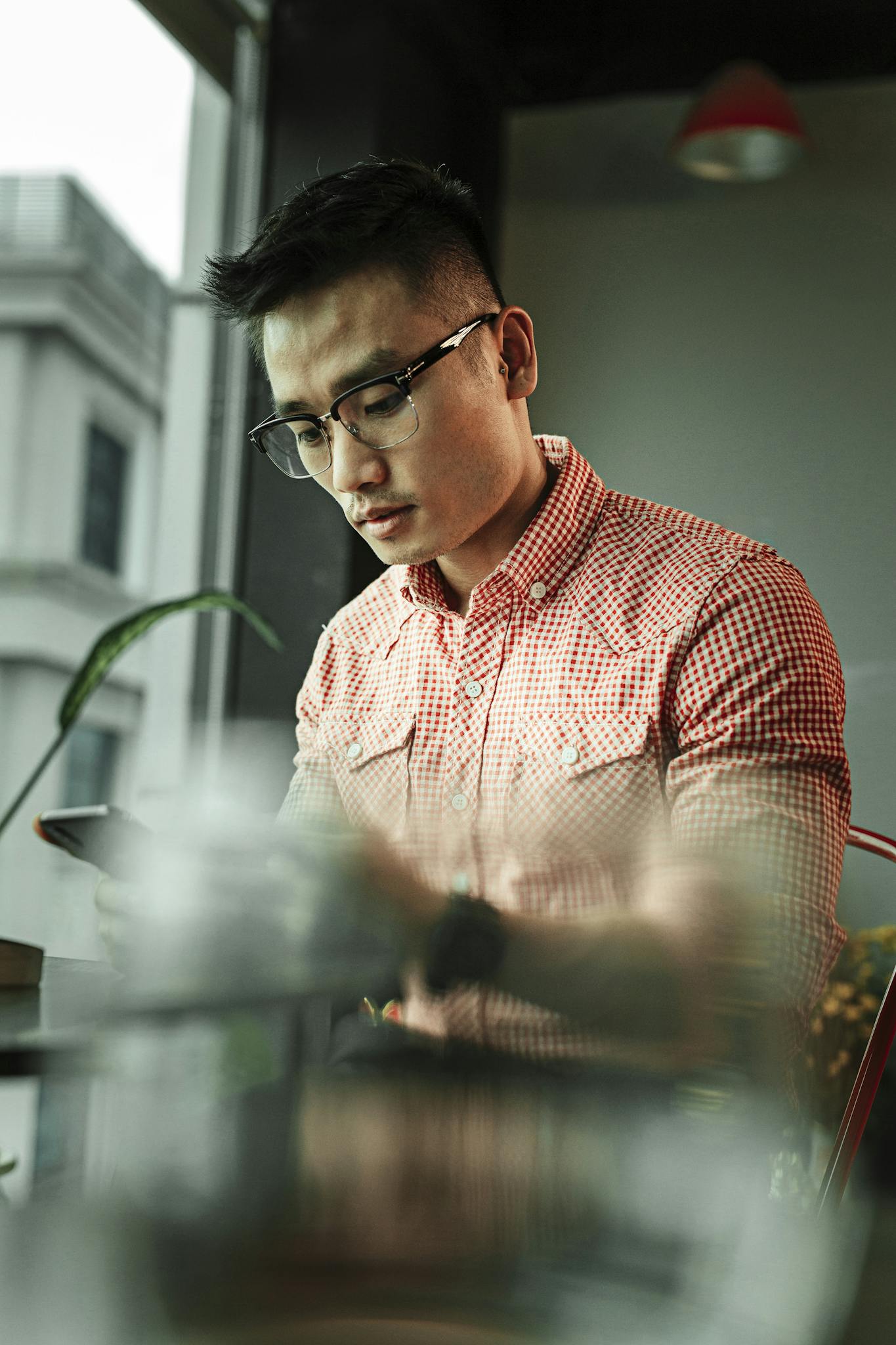 A stylish man with eyeglasses in a checkered shirt using a smartphone by a window. Soft focus effect.