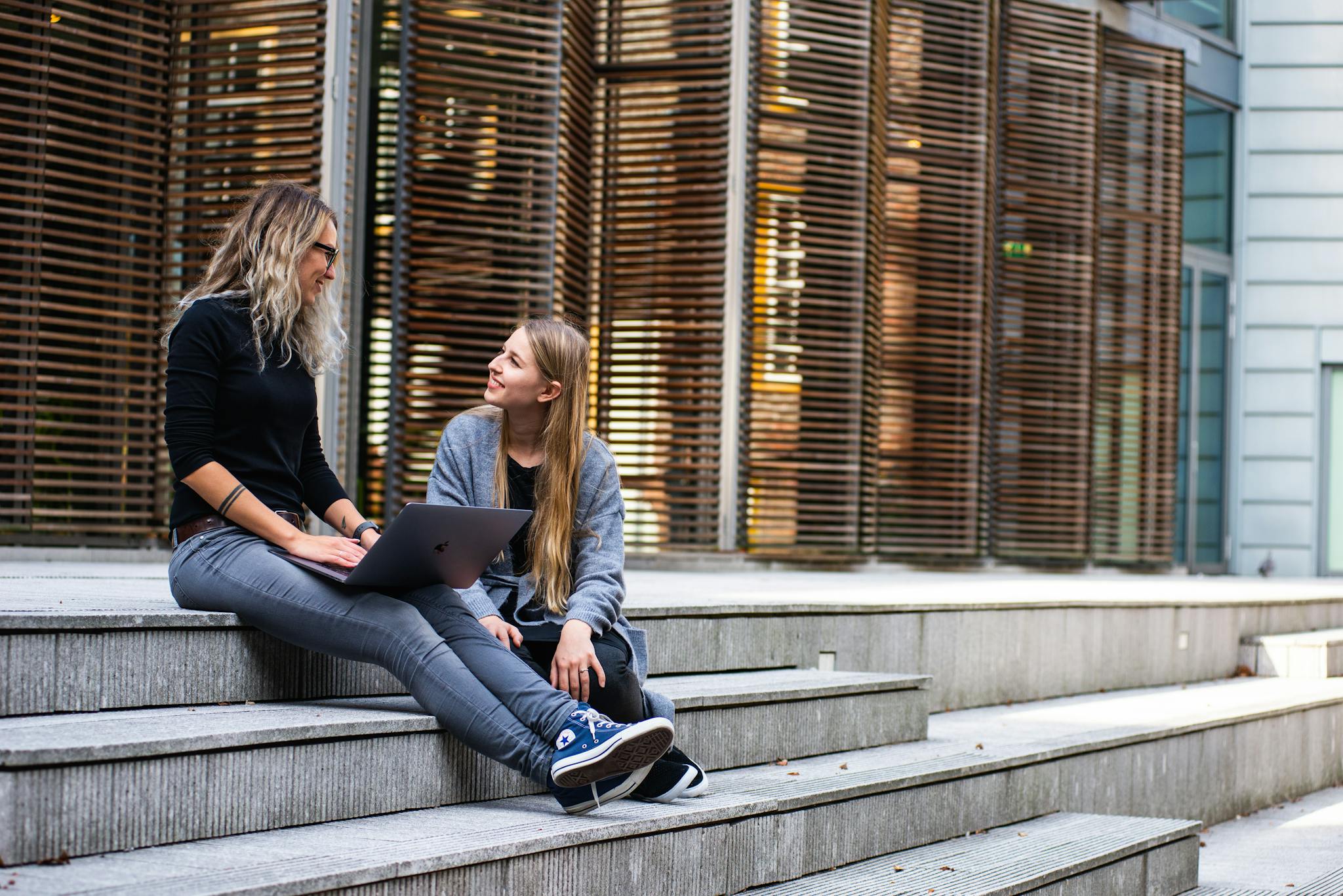 Two women sitting on steps with a laptop, discussing ideas in urban setting.